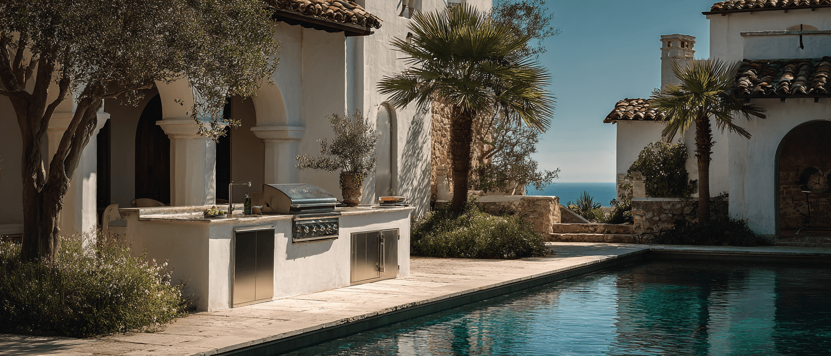 A sun-drenched Mediterranean-style South Bay villa with a built-in grill at the edge of a stone counter, palms framing the courtyard, turquoise pool in the foreground and ocean view beyond.